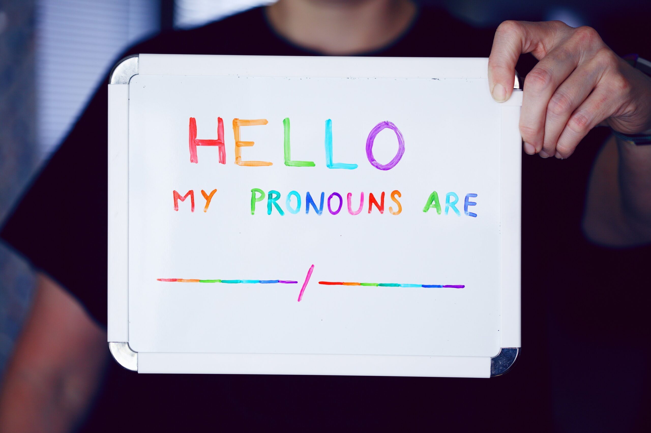 A person you can't see holds up a small whiteboard to show you. They got their hands on the colored whiteboard markers again. In rainbow letters, they've written, "Hello, my pronouns are." After this is a blank line, meant for pronouns. Nothing is written on it. It is also rainbow.