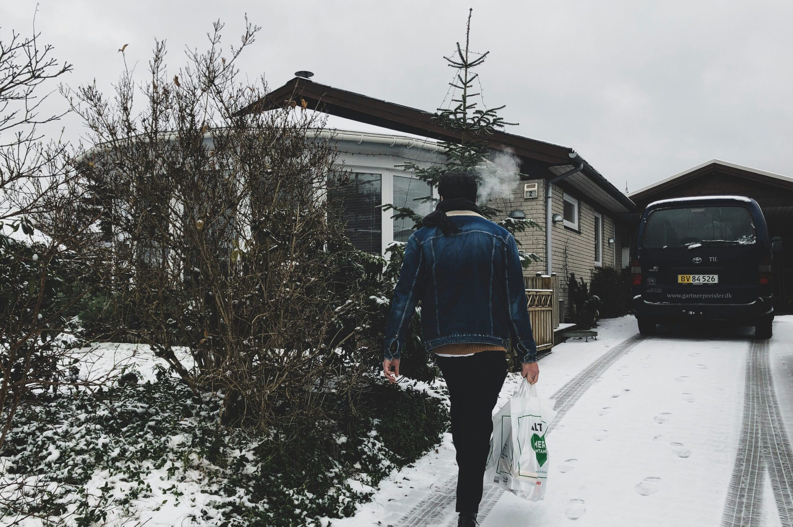 An ordinary person with a great fashion sense carries shopping bags up a snowy driveway towards a house. The house has a really nice sunroom out front. It looks like a ship's bridge. I want a sunroom like that.