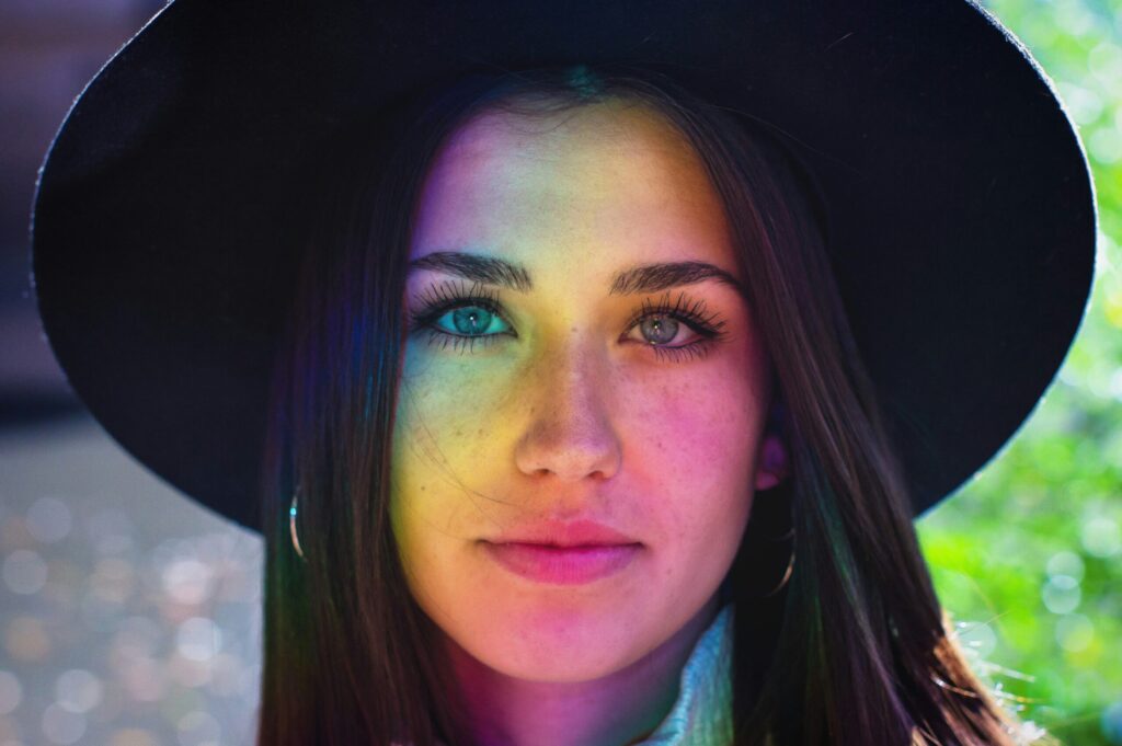 A young woman looks into the camera, smiling faintly. She has long, dark hair and a wide-brimmed hat that the lesbians would love. Rainbow light reflects across her face. It is unclear whether she's standing behind a suncatcher, sneezed into a pot of pixie dust, or just woke up very gay today.